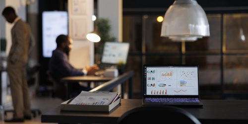 Laptop computer with marketing statistics standing on desk table in startup office, businessmen working overtime at company strategy. African american entrepreneurs brainstorming business ideas