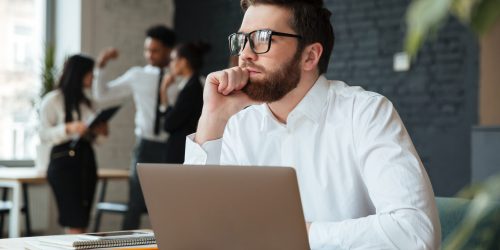 Image of concentrated young caucasian businessman sitting indoors using laptop computer. Looking aside.